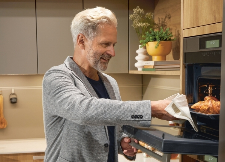 Älterer Mann nimmt mit einem Küchentuch einen Braten aus einem hochgebauten Backofen in einer modernen Küche.