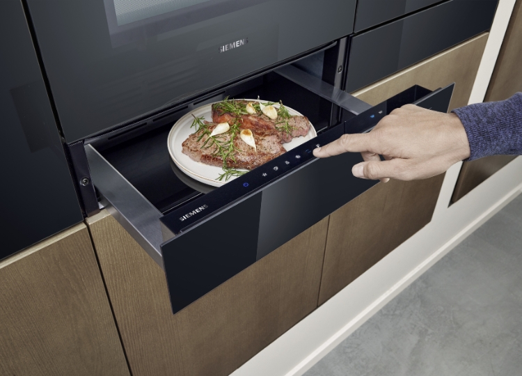 Geöffnete schwarze Wärmeschublade mit einem angerichteten Teller unter einem Einbaubackofen in einer modernen Küche.