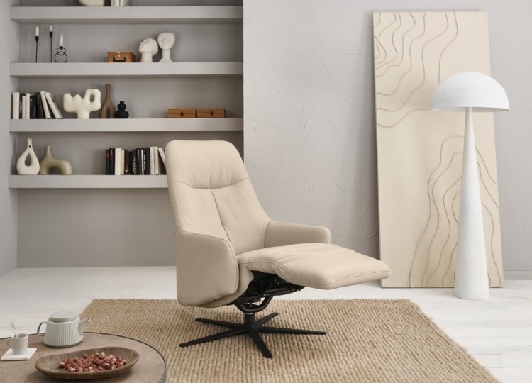 Cremefarbener Relaxsessel mit hoher Rückenlehne, passendem Fußhocker und schwarzem Drehgestell vor einer hellen Regalwand.