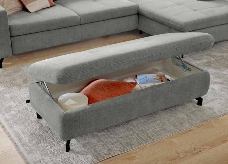 Geöffneter grauer Polsterhocker mit integriertem Stauraum für Decken und Wohnaccessoires vor einem Ecksofa.